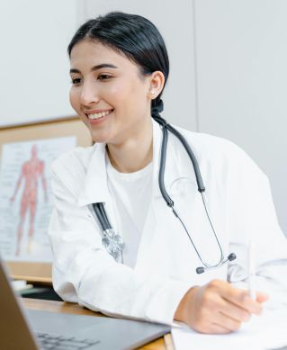 Confident female doctor in a lab coat smiling during an online consultation using a laptop.
