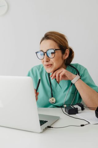 A healthcare professional wearing scrubs and eyeglasses using a laptop with a stethoscope around the neck.